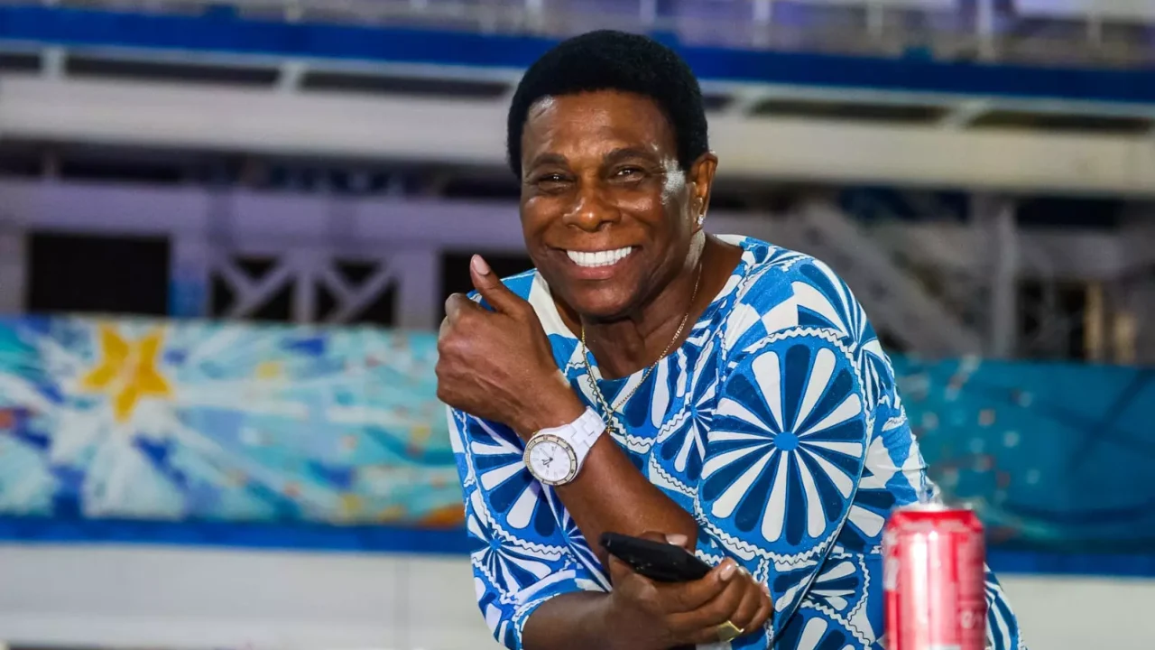 Neguinho da Beija-Flor sorrindo e fazendo joinha durante ensaio da escola de samba, com fantasia azul e branca