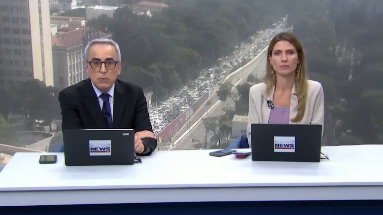 Nelson Gomes e Patricia Rocha apresentam telejornal da BandNews com laptops sobre bancada e trânsito intenso ao fundo