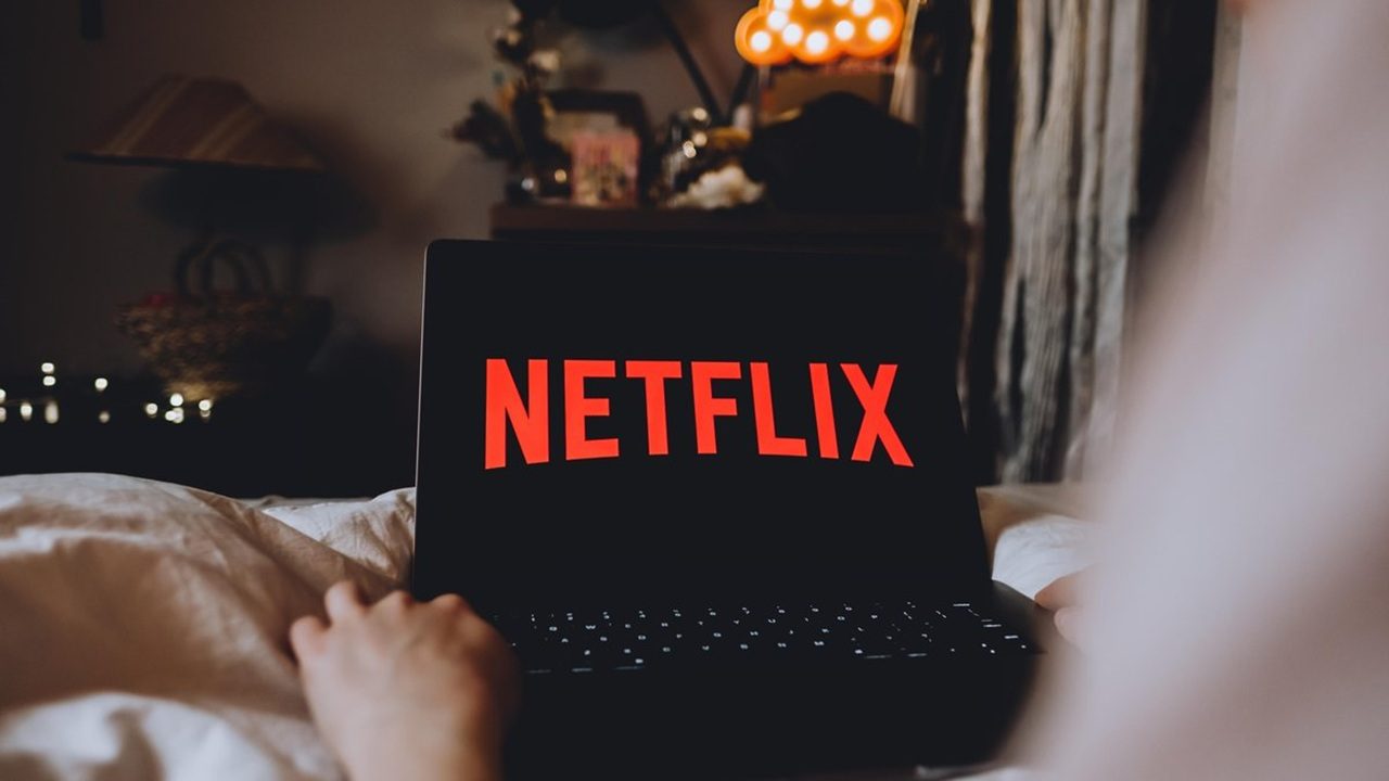 Pessoa deitada em uma cama assiste à tela de um notebook que exibe o logotipo vermelho da Netflix sobre fundo preto. O ambiente ao redor é um quarto com iluminação suave, abajur aceso e objetos decorativos desfocados ao fundo, transmitindo clima aconchegante.