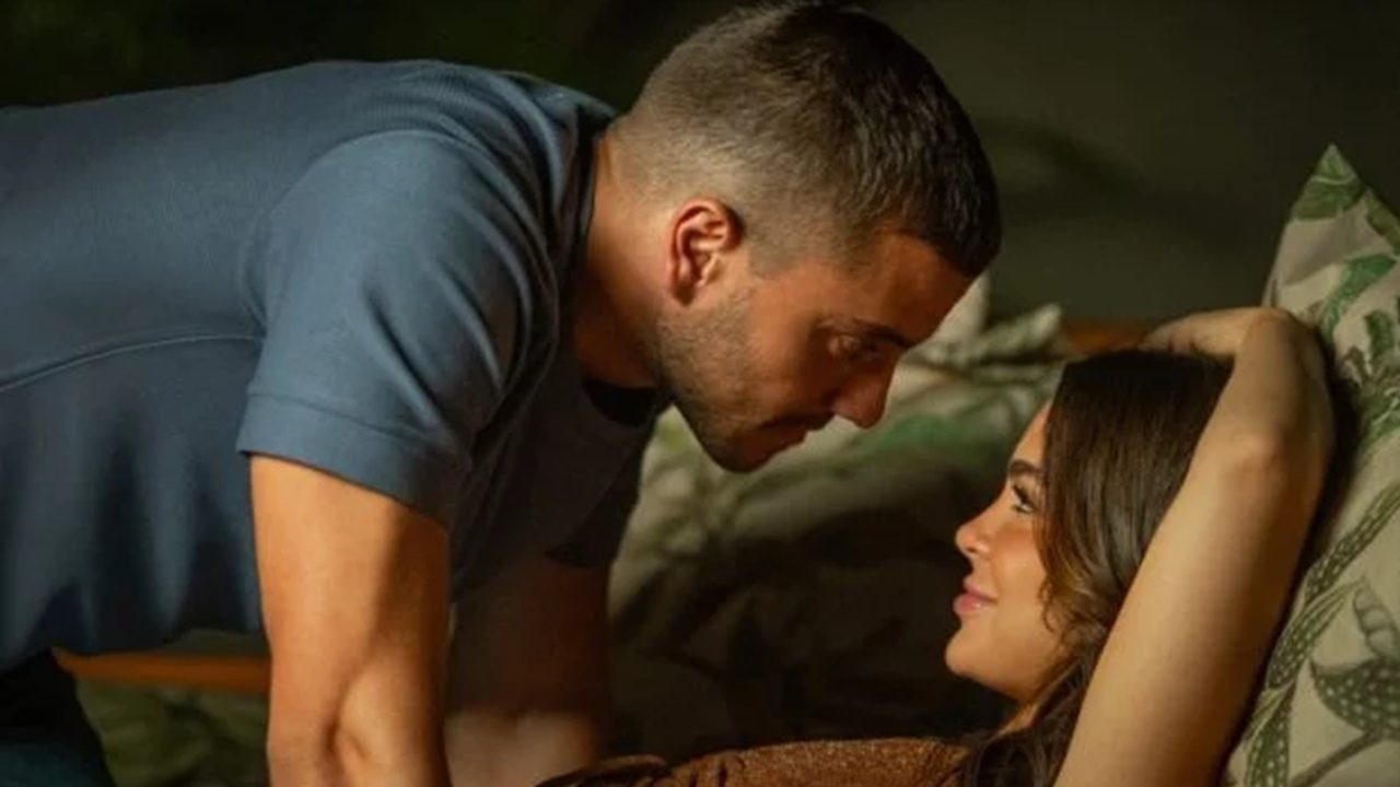 Homem inclinado sobre uma mulher deitada em almofadas trocam olhares próximos e sorridentes em cena de clima romântico e intimista.