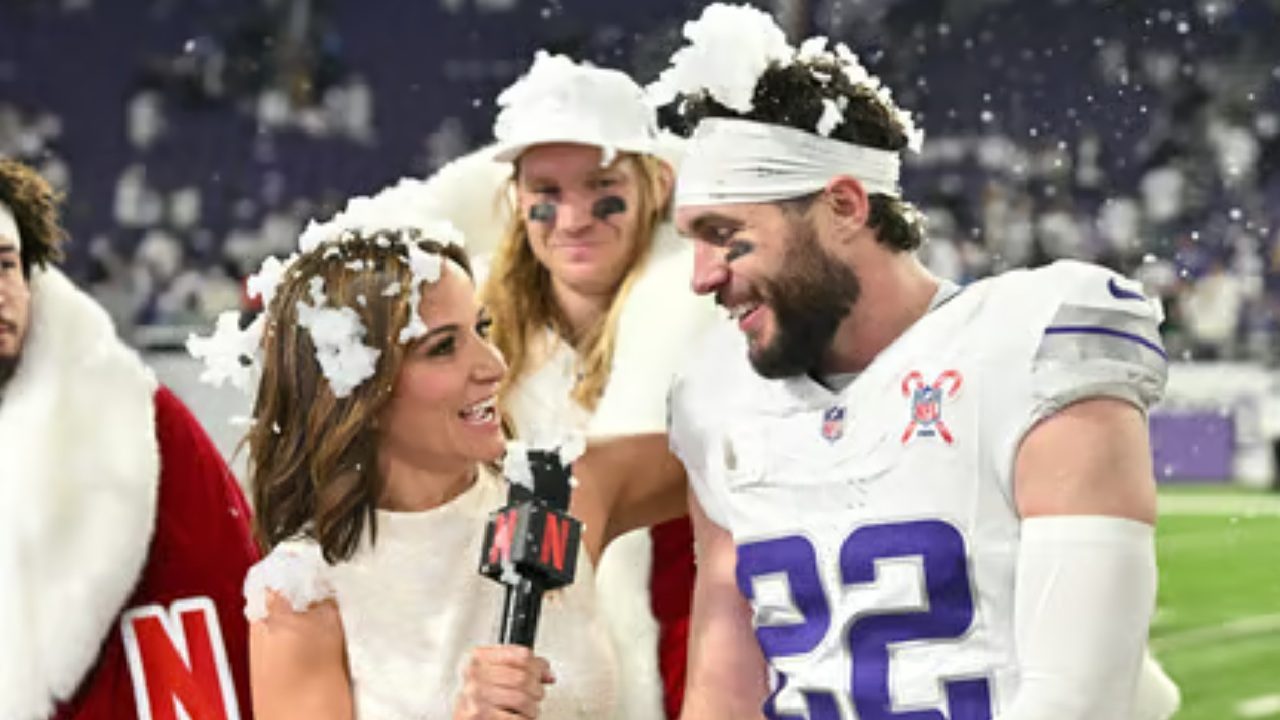 Mulher com microfone da Netflix entrevista jogador de futebol americano com uniforme branco e número 22, ambos cobertos por flocos de neve falsa enquanto sorriem dentro de um estádio iluminado, com outras pessoas fantasiadas e torcedores desfocados ao fundo.