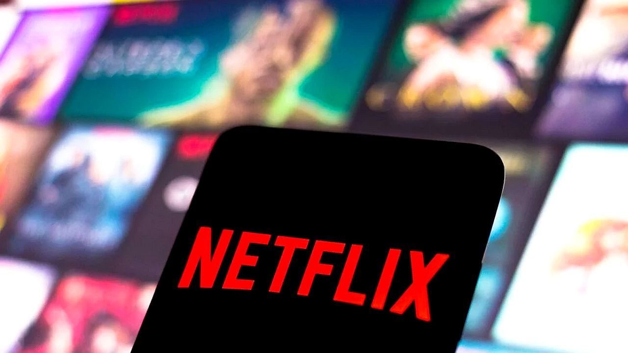 Logotipo da Netflix exibido em tela de celular com catálogo de filmes e séries desfocado ao fundo
