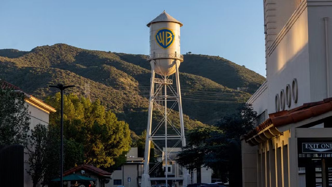 Caixa d'água metálica com o logotipo da Warner Bros. em destaque, erguida sobre uma torre alta no estúdio da empresa. Ao fundo, montanhas iluminadas pela luz do fim de tarde e prédios do complexo cinematográfico
