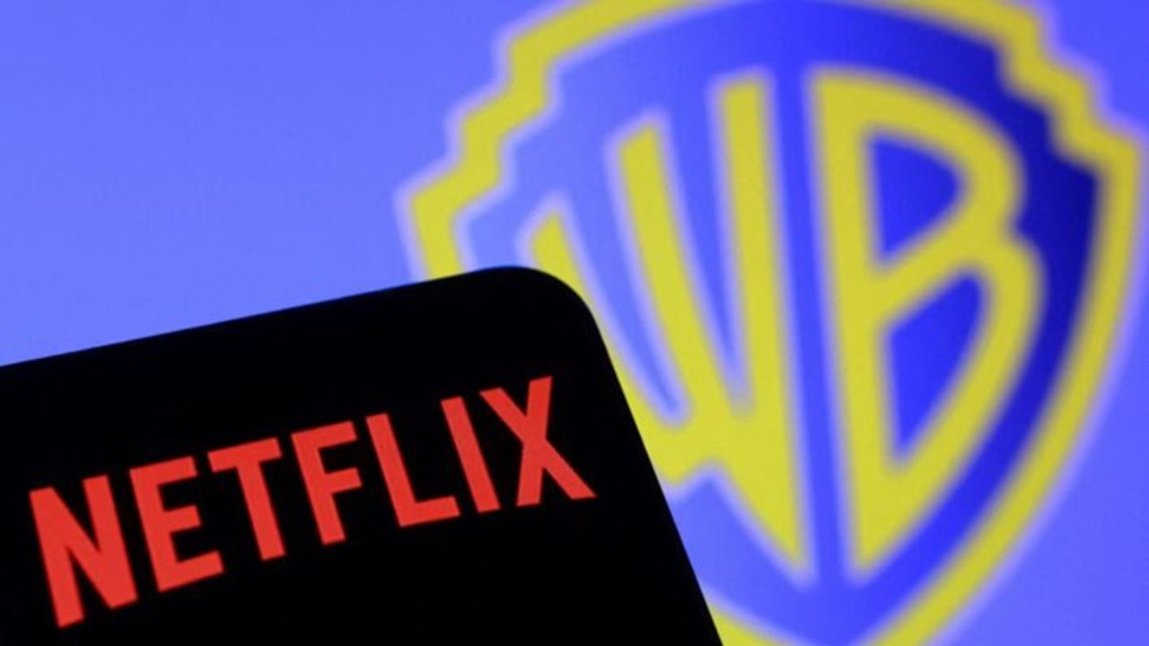 Tela mostra em primeiro plano o canto de um dispositivo escuro com o logotipo vermelho da Netflix em letras maiúsculas enquanto ao fundo aparece desfocado o escudo da Warner Bros em azul e amarelo ocupando grande parte da imagem sugerindo relação ou negociação entre as duas empresas de entretenimento em contexto corporativo, ou midiático