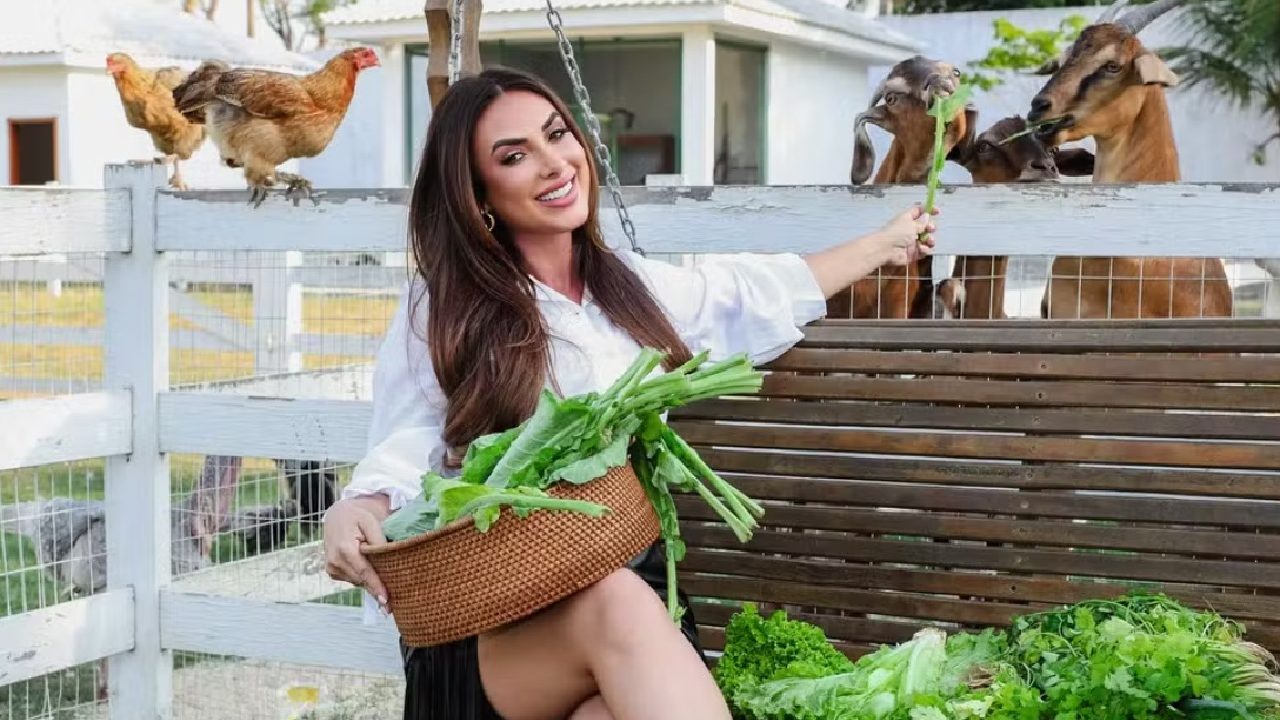 Nicole Bahls sentada em balanço de madeira sorri enquanto segura uma cesta com hortaliças e alimenta cabras em um cercado; ao fundo há galinhas e construções rurais.