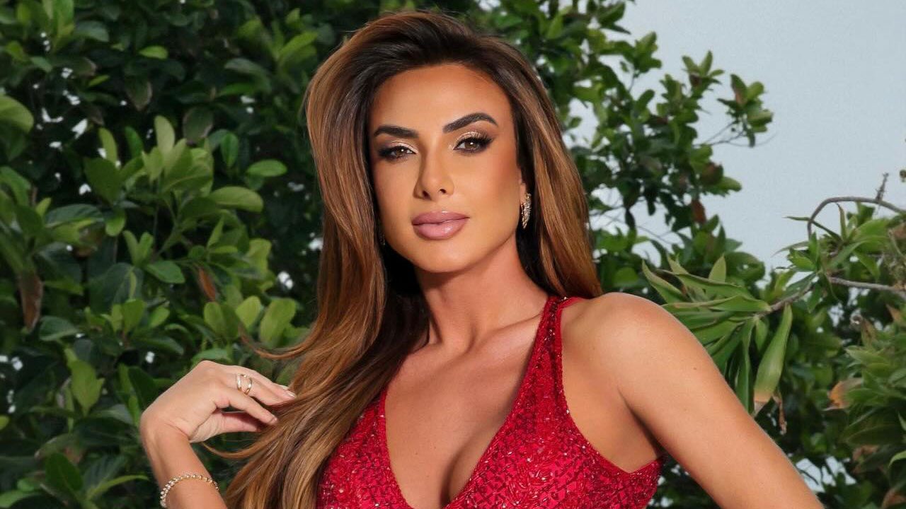 Nicole Bahls usa vestido vermelho brilhante e posa com a mão nos cabelos soltos em cenário externo com folhagens verdes ao fundo