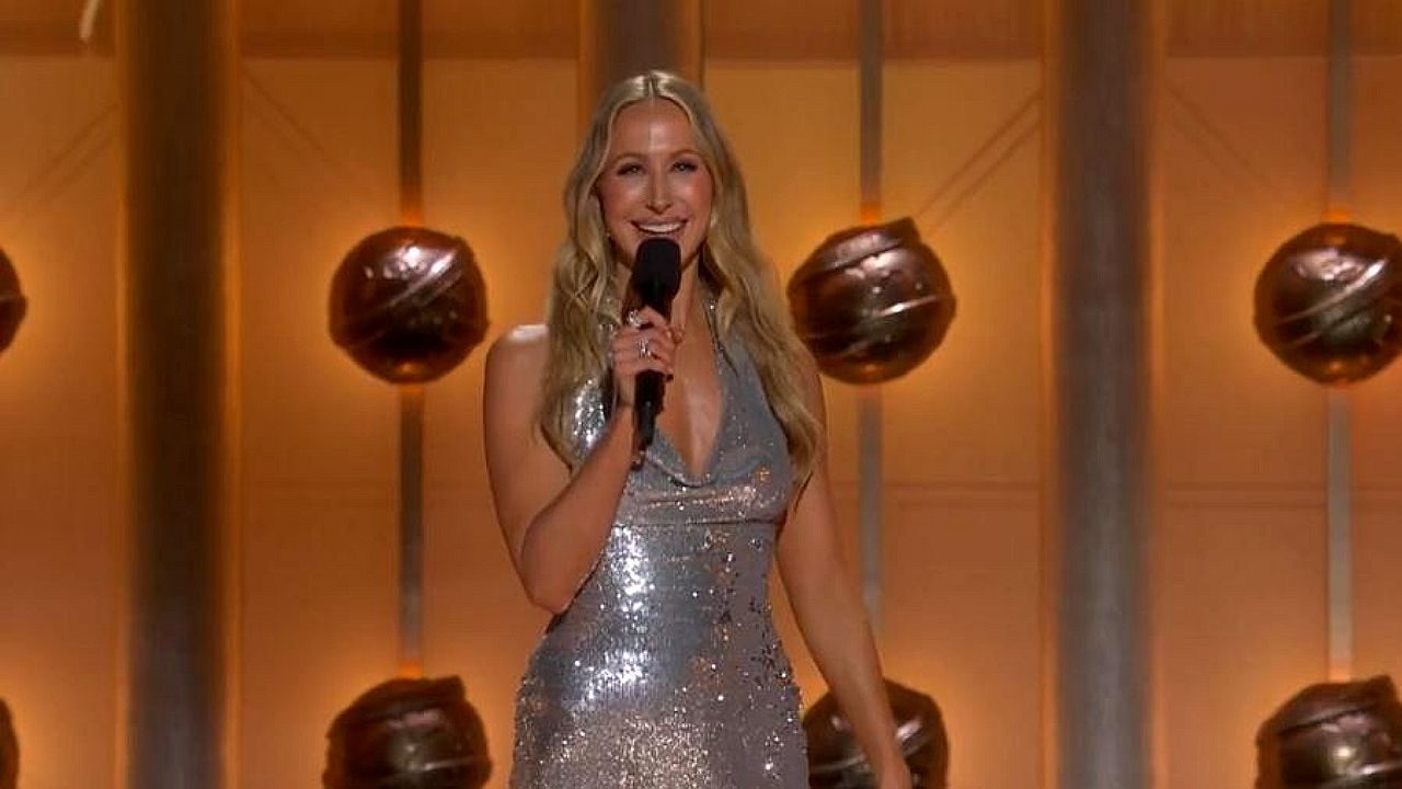 Apresentadora de vestido prateado segura microfone no palco durante premiação televisiva