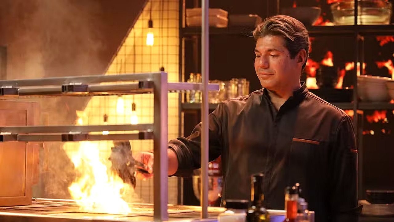 Chef manipula grelha com chamas altas em cozinha profissional durante preparo de prato em programa culinário