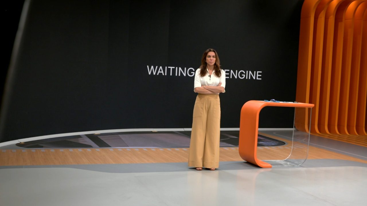 Apresentadora fica de braços cruzados ao lado de bancada laranja com tela exibindo mensagem waiting engine no estúdio do Bom Dia Brasil