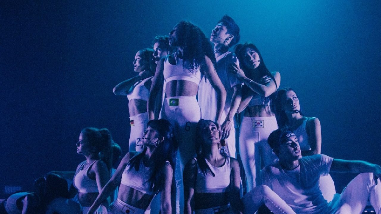 Grupo de dançarinos no palco faz pose coreografada sob iluminação azul; todos vestem roupas brancas esportivas com bandeiras de diferentes países, transmitindo energia e unidade em apresentação artística