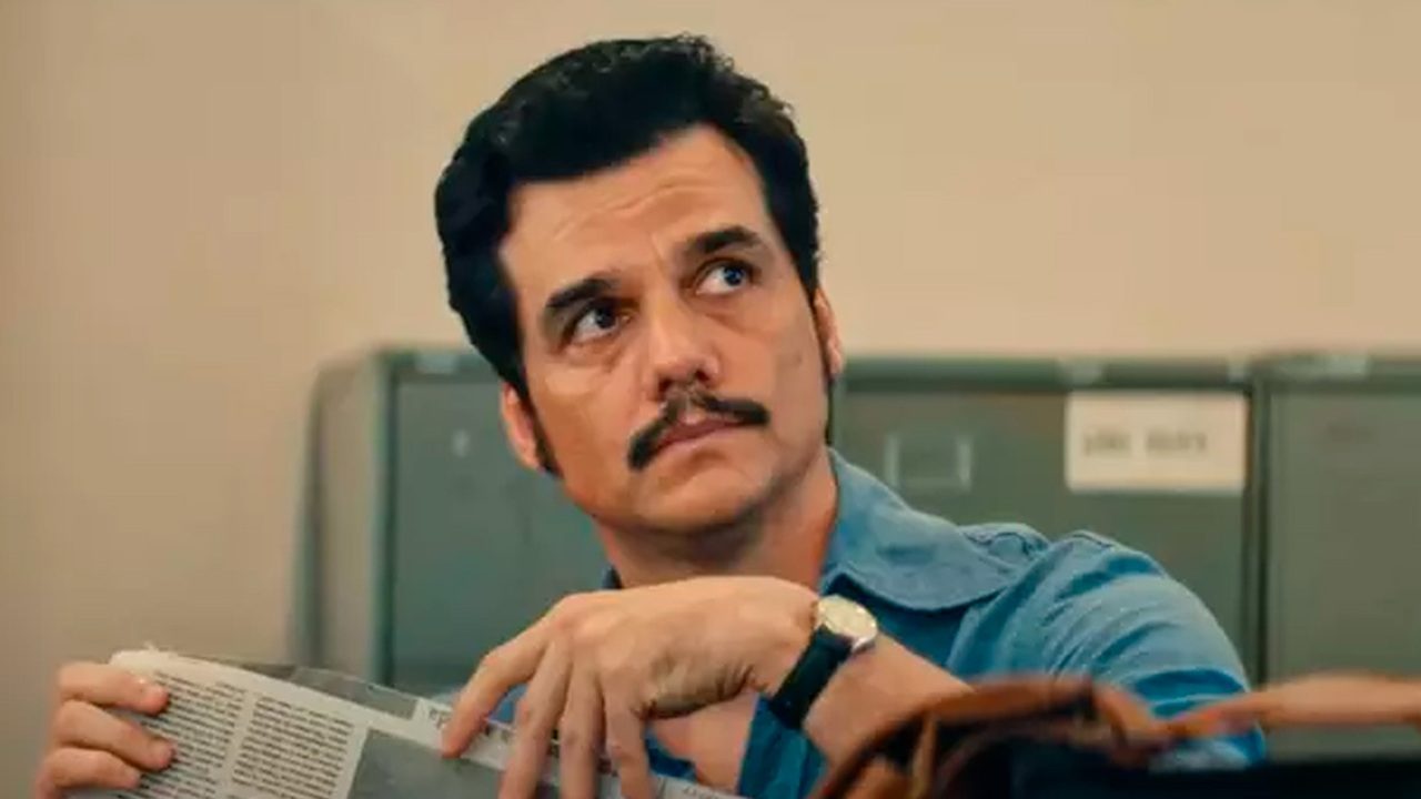 Homem de bigode e camisa azul segura jornal e olha para o lado em cena ambientada em escritório