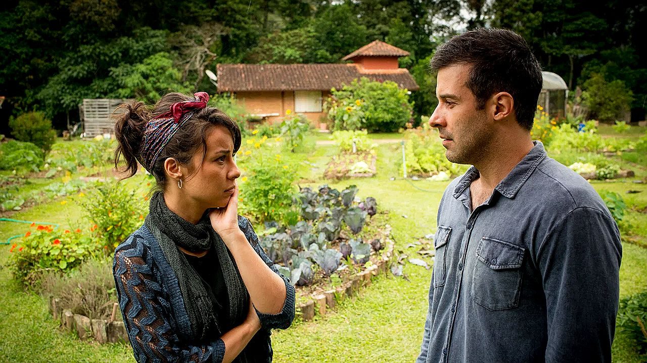 Mulher e homem conversam em horta ao ar livre com casa ao fundo em cena de novela