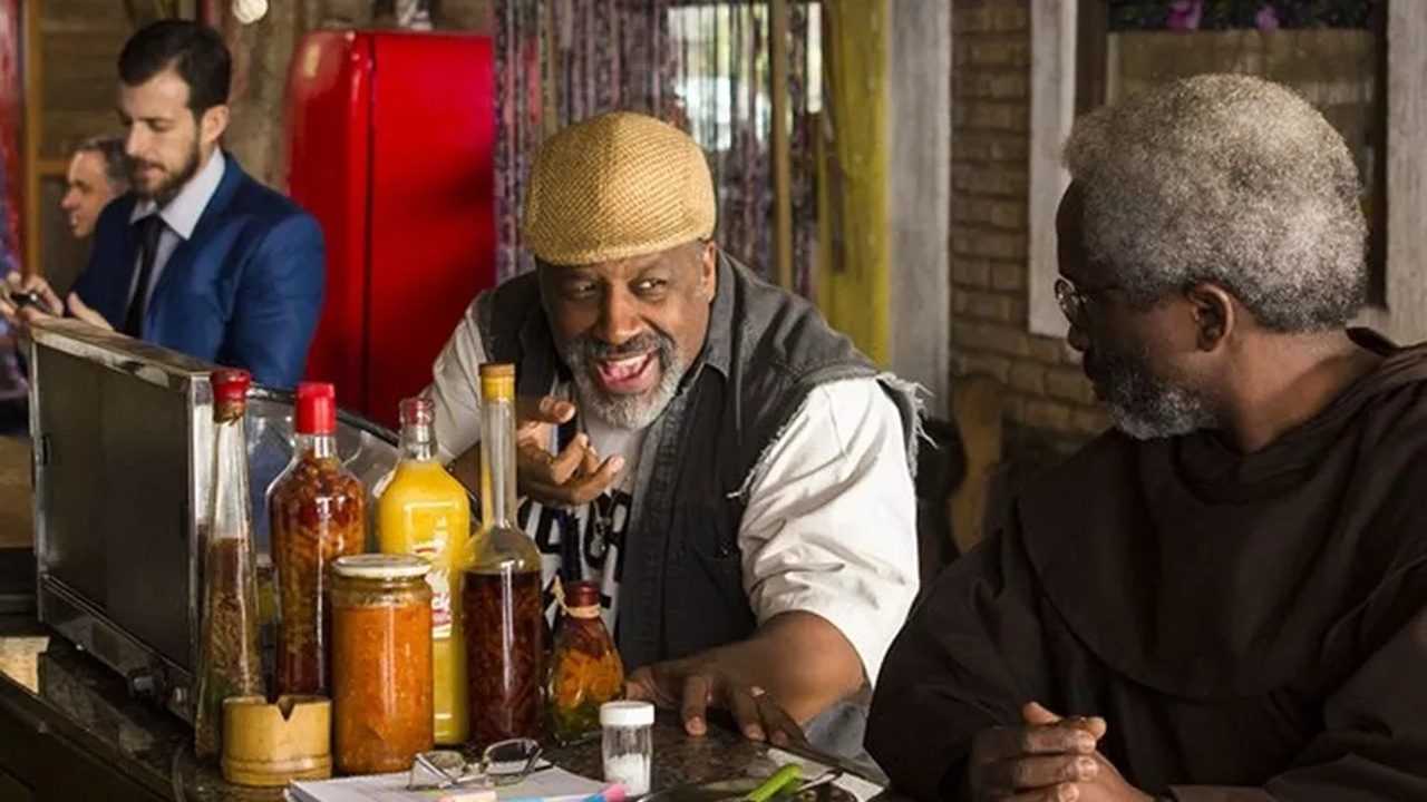 Dois homens conversam animadamente em um balcão com várias garrafas e potes à frente; um deles usa boina clara e sorri amplamente, enquanto o outro, de barba grisalha e roupas escuras, o observa com atenção.