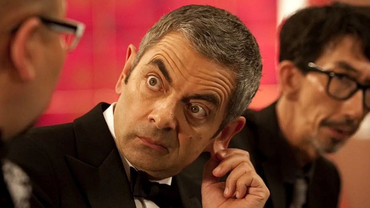 Homem de terno preto e gravata borboleta está sentado à mesa em ambiente elegante, olhando de forma intensa e arregalada para outra pessoa ao seu lado. Com expressão surpresa e levemente desconfiada, ele leva a mão à orelha, enquanto ao fundo outro homem de óculos aparece desfocado. A cena sugere momento cômico ou de tensão em evento formal.