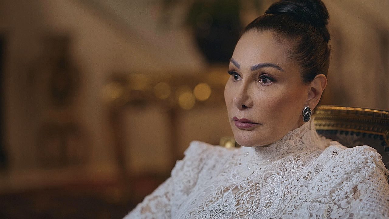 Mulher com cabelo preso e vestido branco rendado observa atentamente em cena de entrevista em ambiente elegante