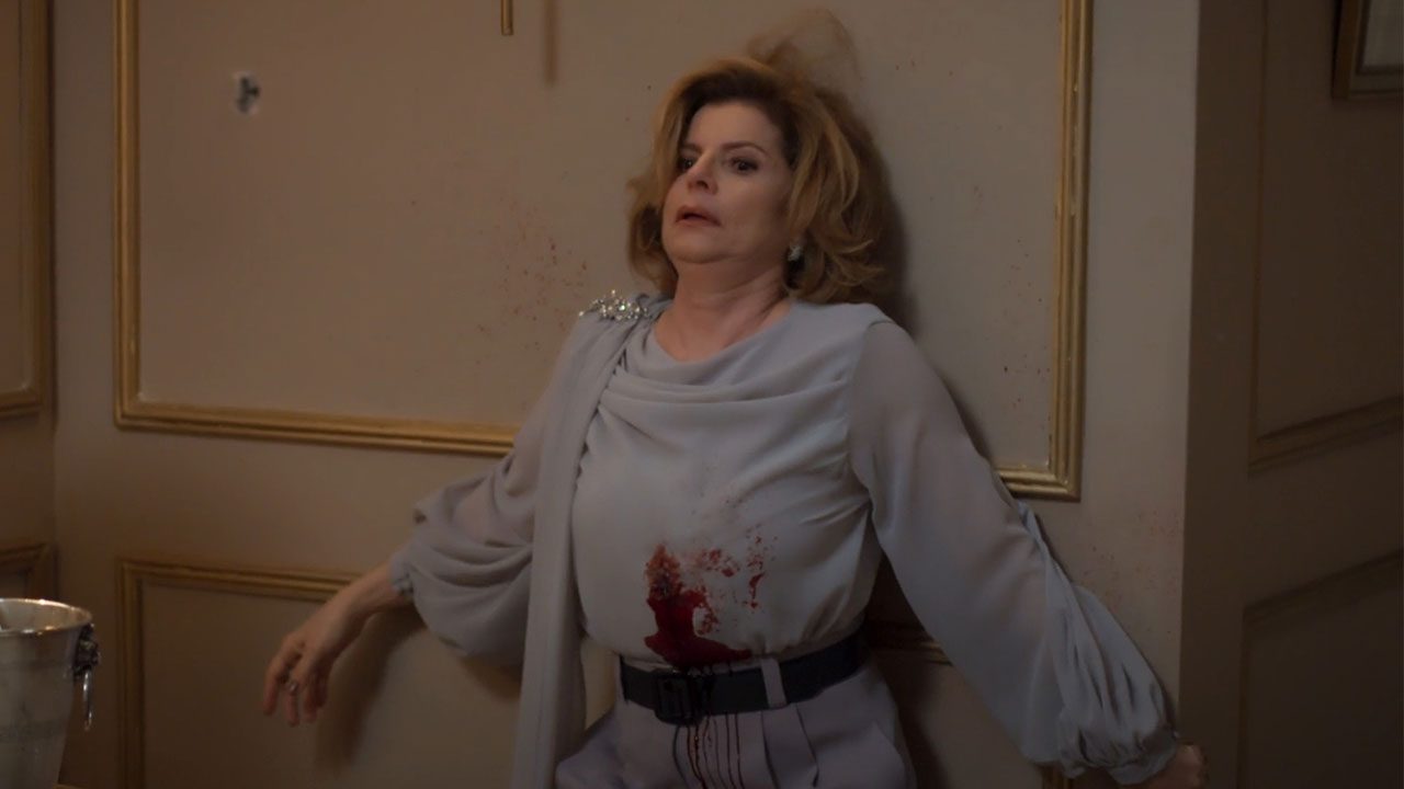 Débora Bloch como Odete Roitman na cena de sua morte em Vale Tudo; a vilã está caída contra a parede com a roupa ensanguentada