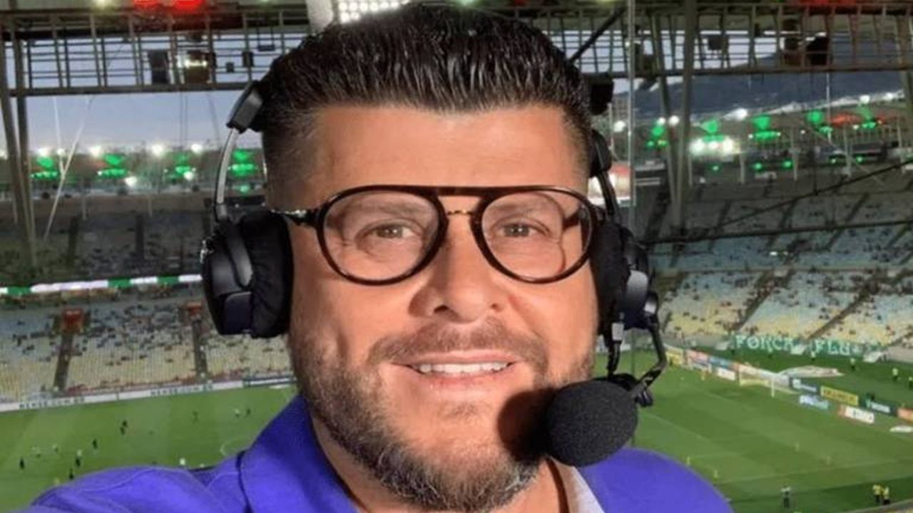 A imagem mostra um comentarista esportivo com fones de ouvido e microfone, provavelmente durante uma transmissão ao vivo de futebol. Ao fundo, é possível ver um estádio iluminado e o campo de jogo com jogadores se aquecendo.