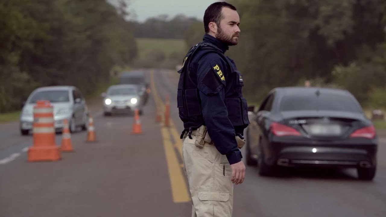Agente de polícia rodoviária, usando uniforme azul escuro com colete tático e calça bege, está parado no meio de uma rodovia durante uma operação de fiscalização. Cones laranja delimitam a área, enquanto carros se aproximam lentamente pela pista. A cena ocorre em um trecho cercado por vegetação