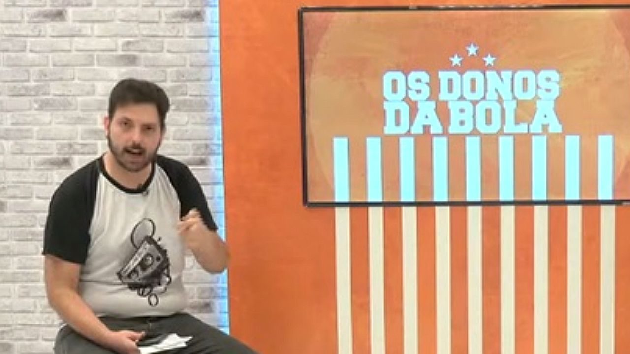 Apresentador comenta futebol ao lado de tela com logotipo do programa Os Donos da Bola