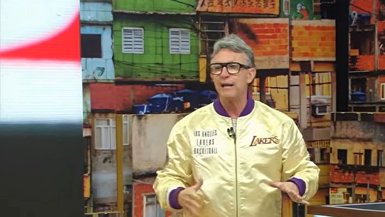 Apresentador Neto comenta no Os Donos da Bola diante de telão com imagem de comunidade ao fundo