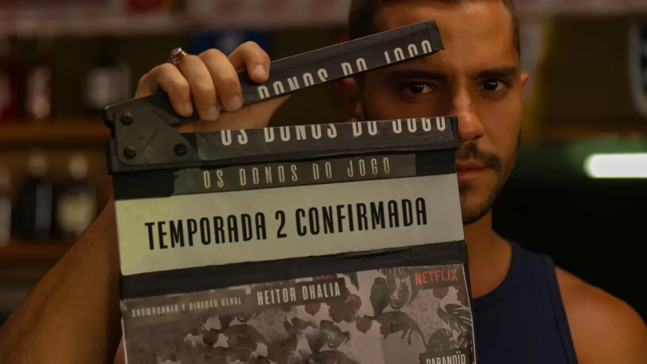 Homem segura uma claquete de filmagem com a inscrição “Os Donos do Jogo — Temporada 2 Confirmada”. Abaixo, aparecem os créditos de direção de Heitor Dhalia e o logotipo da Netflix. A imagem indica o anúncio oficial da segunda temporada da série, com o ator olhando diretamente para a câmera em ambiente interno com iluminação quente