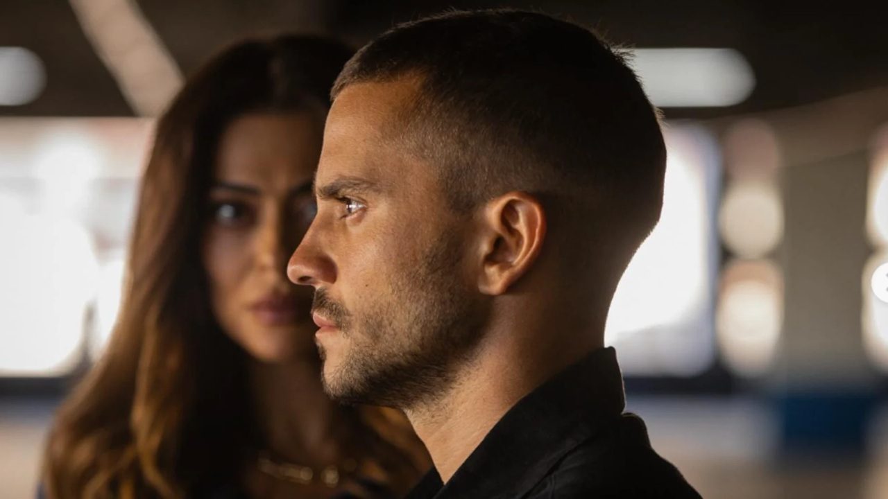 Homem de perfil encara à frente com mulher desfocada ao fundo em cena tensa de série policial