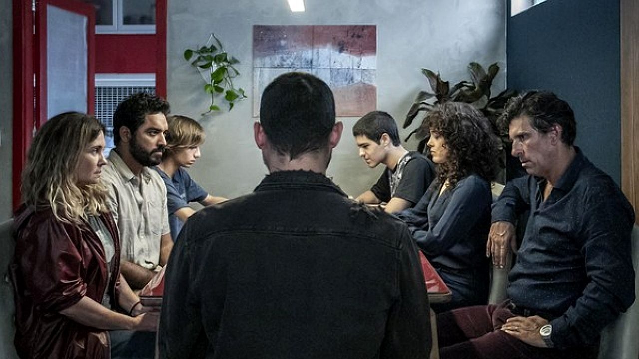 Um grupo de pessoas está reunido ao redor de uma mesa em um ambiente interno, com clima tenso e silencioso. No centro, um homem aparece de costas, enquanto os demais — homens, mulheres e jovens — estão sentados ao redor, com expressões sérias e introspectivas, muitos olhando para baixo. O espaço tem decoração simples, com plantas e um quadro na parede. A iluminação é suave, reforçando o tom dramático da cena, que sugere uma conversa difícil ou momento de confronto.