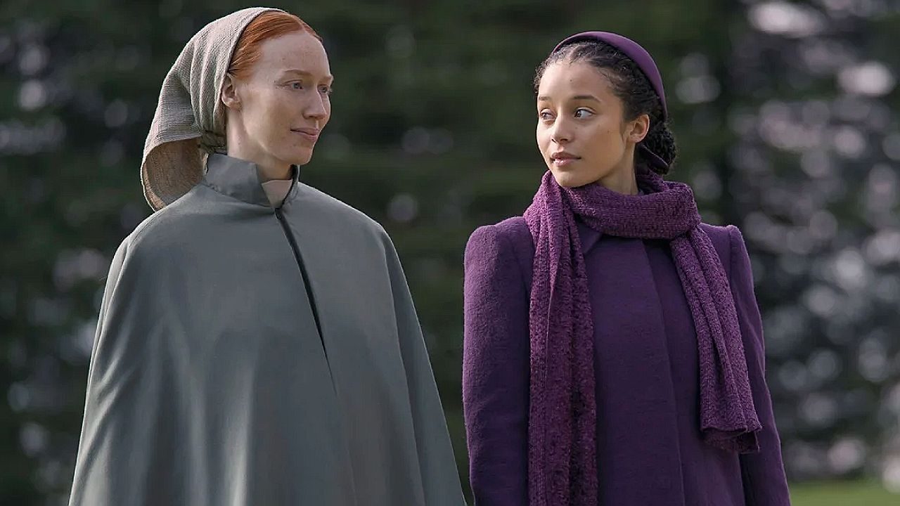 Duas mulheres caminham lado a lado e trocam olhares em cena de série ao ar livre com árvores ao fundo