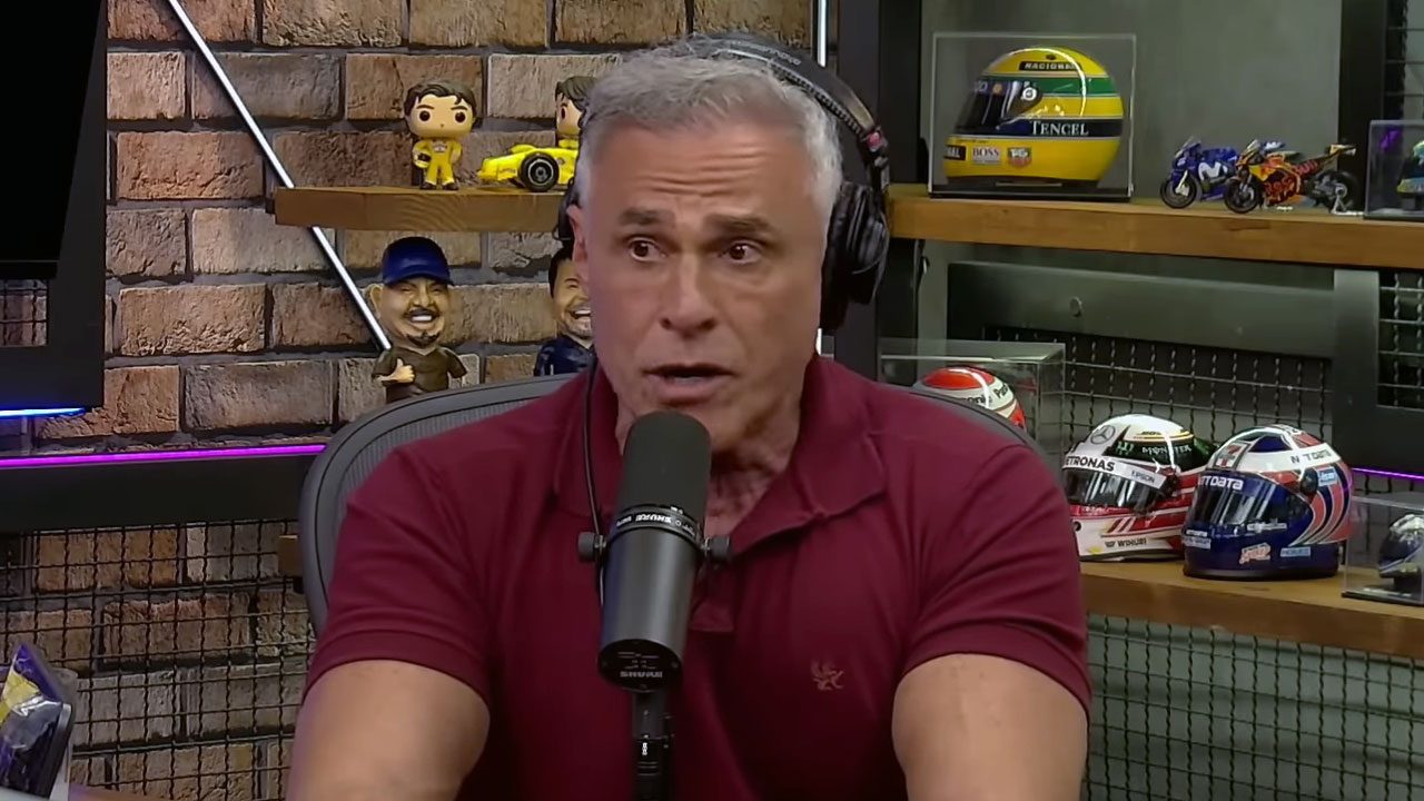 Oscar Magrini falando ao microfone durante participação em podcast com fones de ouvido e mini capacetes ao fundo