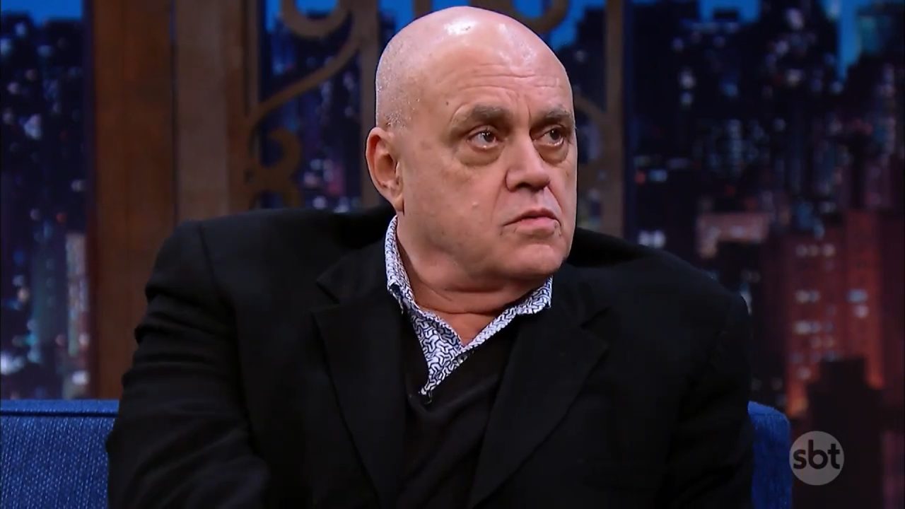 A imagem mostra Oscar Maroni vestindo terno preto e camisa estampada, sentado em um sofá azul durante uma entrevista no The Noite, um programa de televisão do SBT. O cenário ao fundo exibe uma cidade iluminada, característica de programas noturnos de talk show