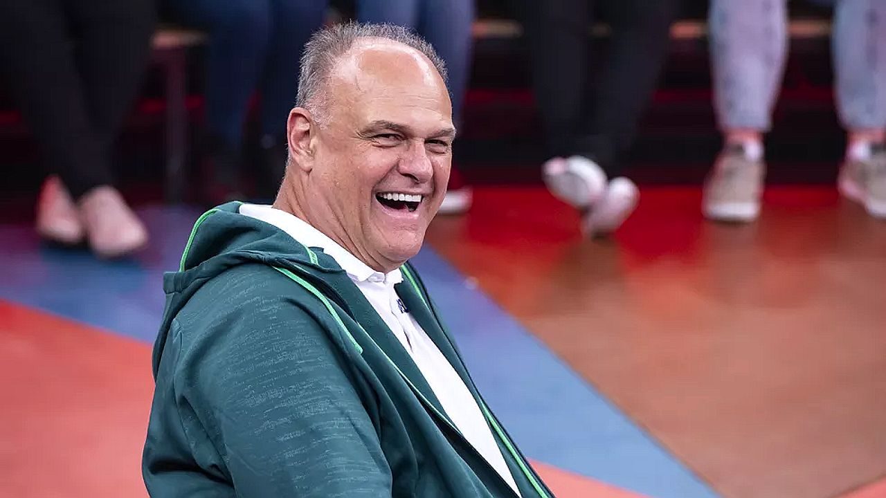 Homem ri sentado em estúdio de TV com plateia ao fundo durante programa de auditório