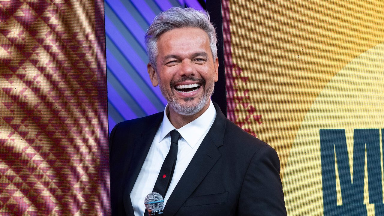 Otaviano Costa sorrindo de terno preto e gravata segurando microfone no palco do Melhor da Noite da Band