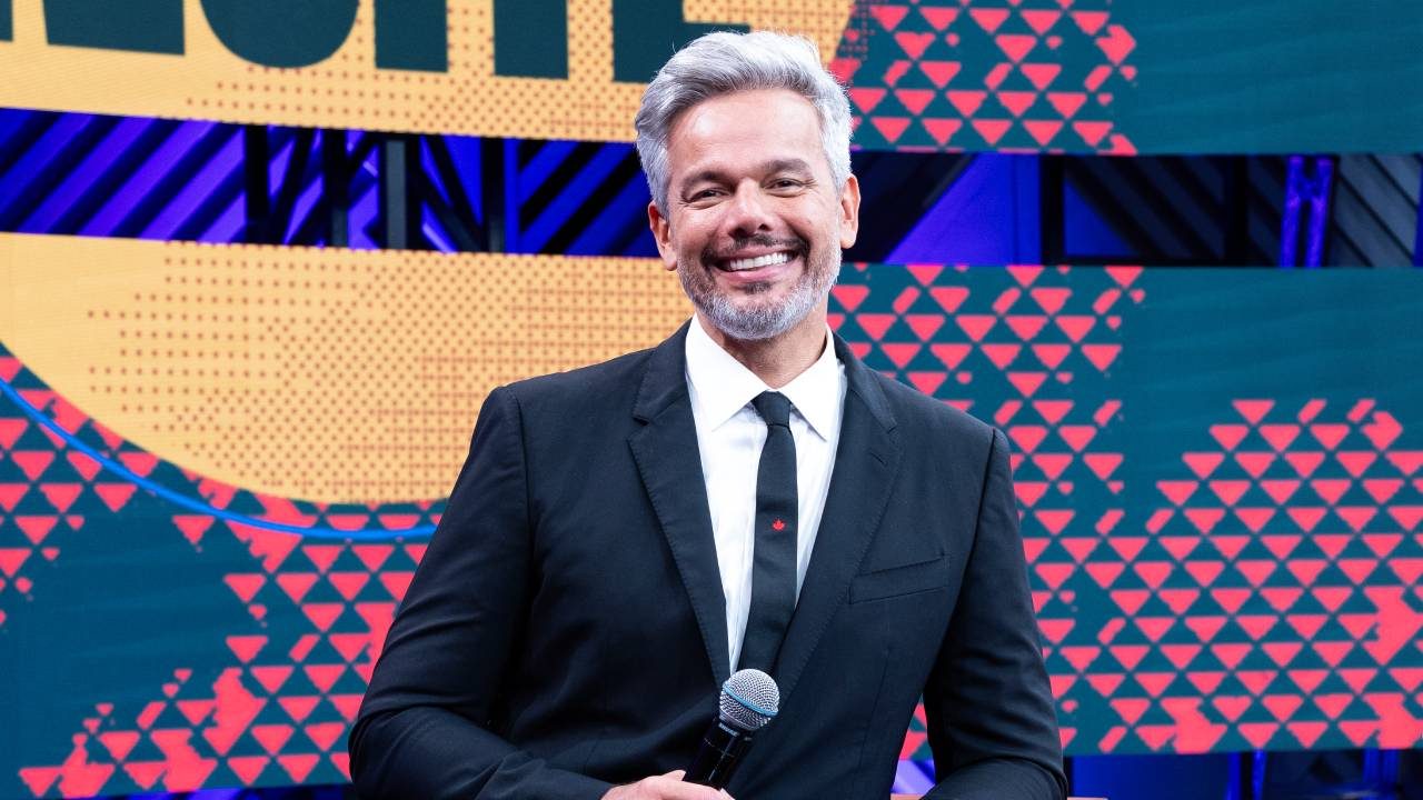Otaviano Costa sorri de terno preto e gravata segurando microfone no cenário do programa Melhor da Noite da Band