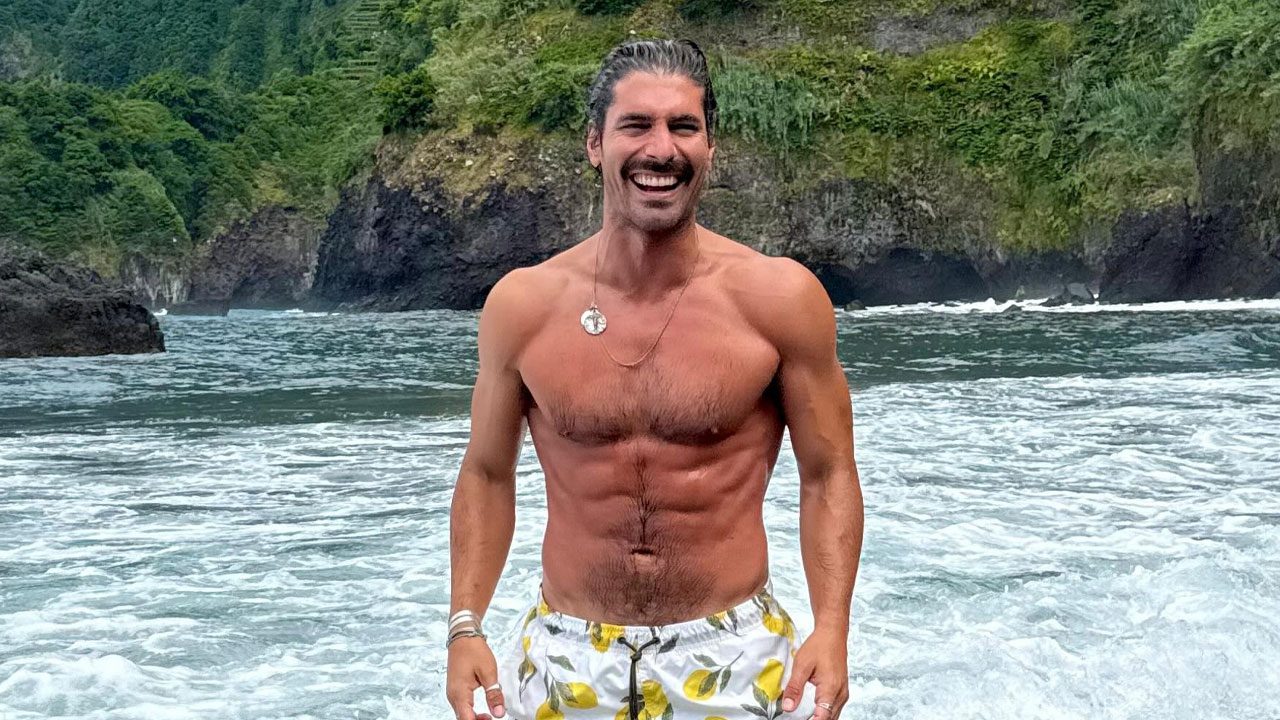 Pablo García sorri de bermuda estampada enquanto posa sem camisa em praia com mar agitado e falésias ao fundo