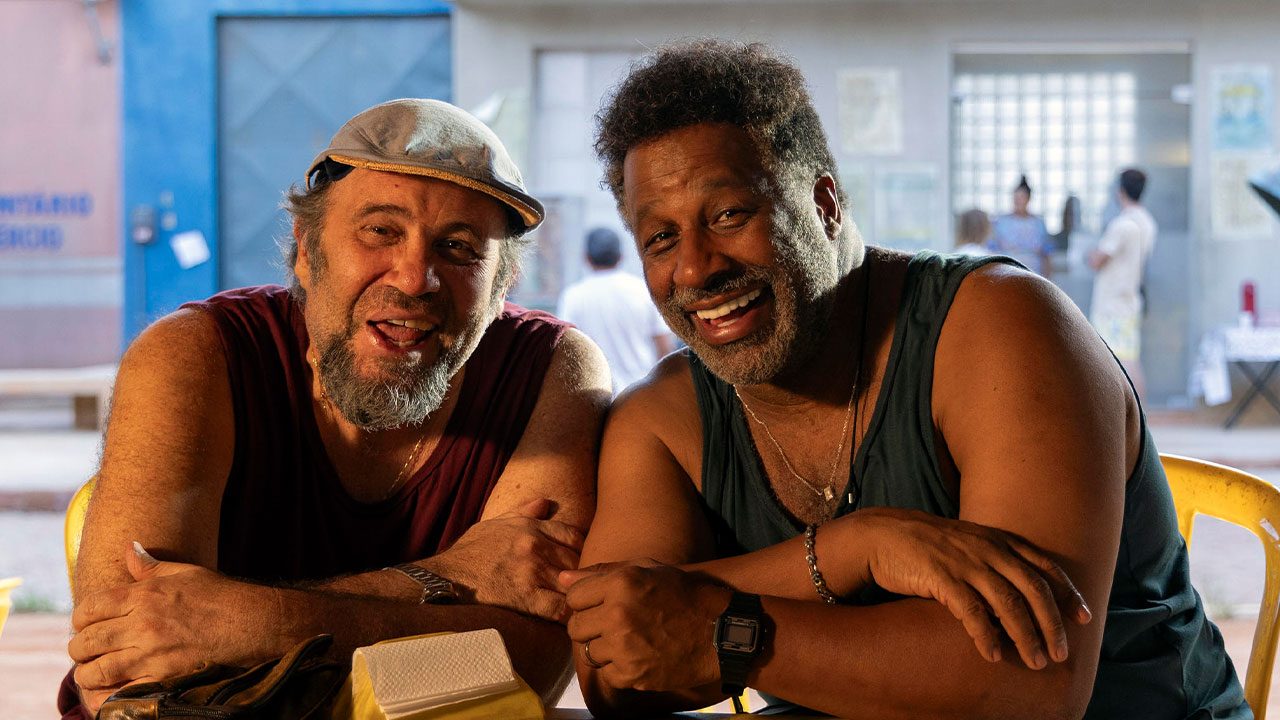 Pablo (Otavio Muller) e Luisão (Ailton Graça) sorrindo sentados à mesa em cena da série Pablo e Luisão