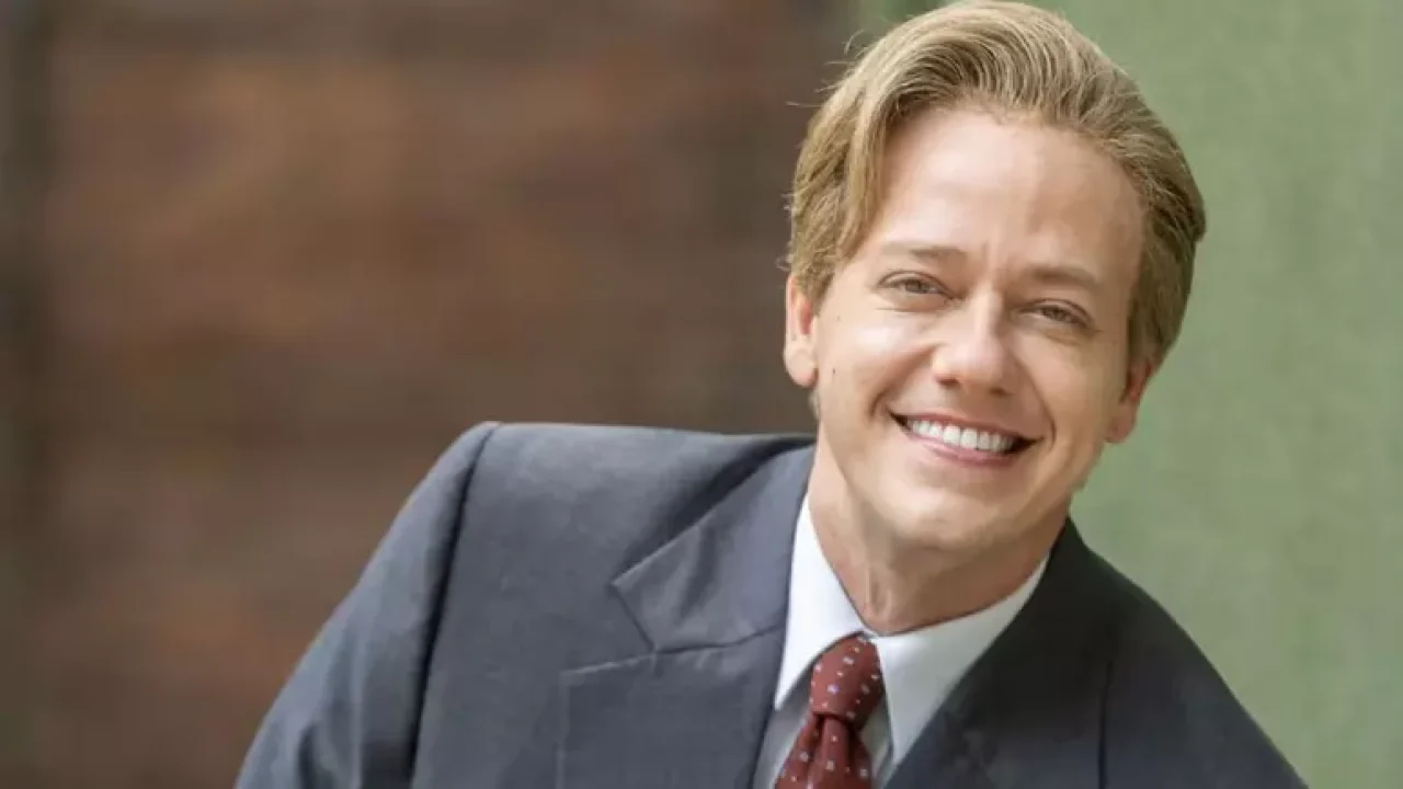 Pablo Sanábio sorrindo caracterizado com terno e gravata em cena da novela Garota do Momento