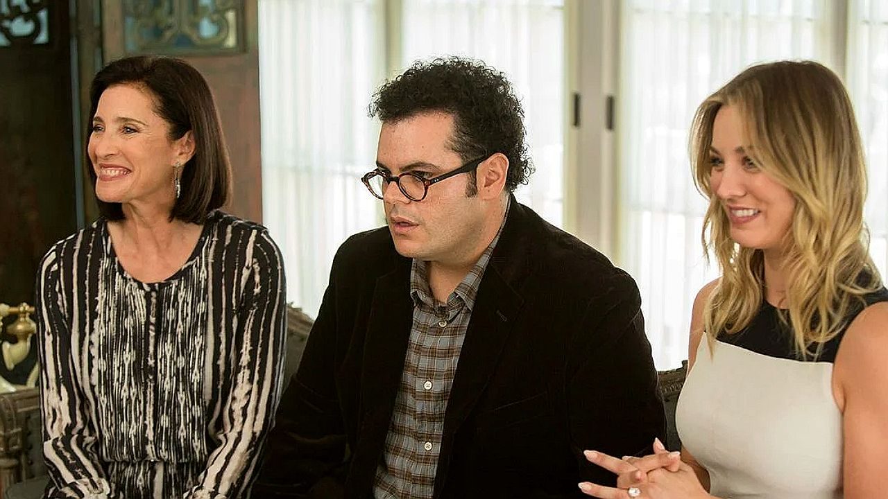 Texto Alternativo disse: Três pessoas conversam e sorriem sentadas à mesa em ambiente elegante em cena de filme