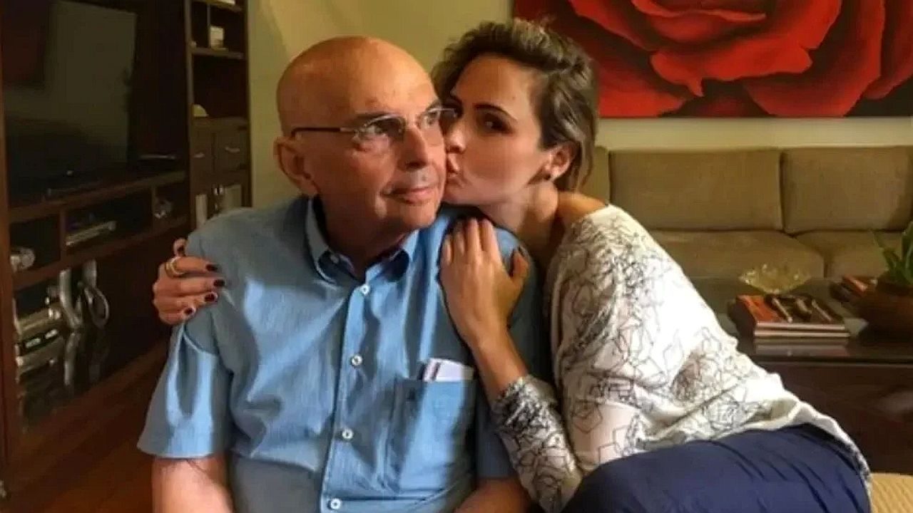 Mulher abraça e beija homem idoso no rosto em sala de estar com sofá e quadro ao fundo