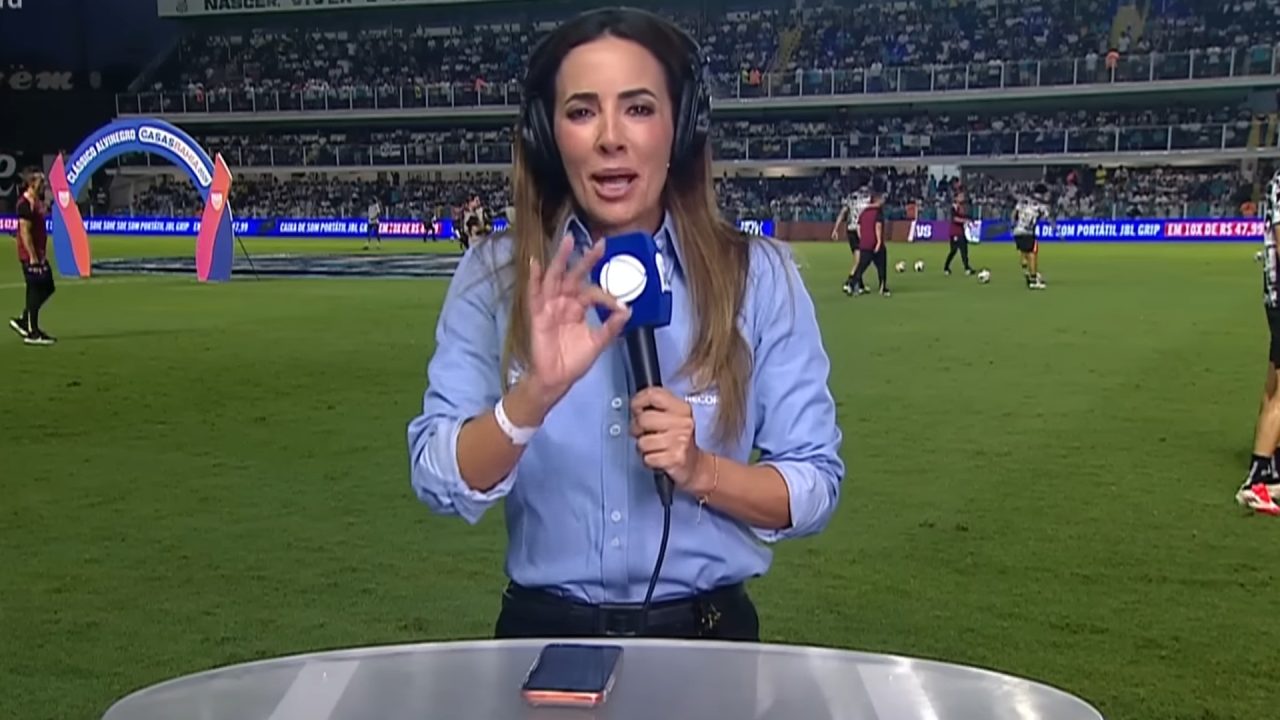 Paloma Tocci, da Record, faz entrada ao vivo direto da Vila Belmiro, segurando microfone com o logotipo da emissora. Ela usa fones de ouvido e camisa azul, enquanto o campo e jogadores do Santos e do Corinthians aquecendo aparecem ao fundo