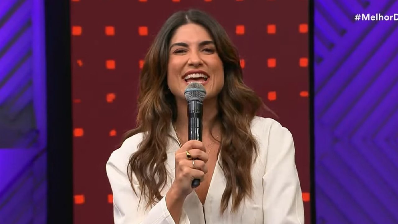 Mulher sorridente segura um microfone enquanto fala em um estúdio colorido, com fundo em tons de roxo e vermelho decorado com quadrados e padrões geométricos. Ela tem cabelos castanhos ondulados e veste uma blusa clara.