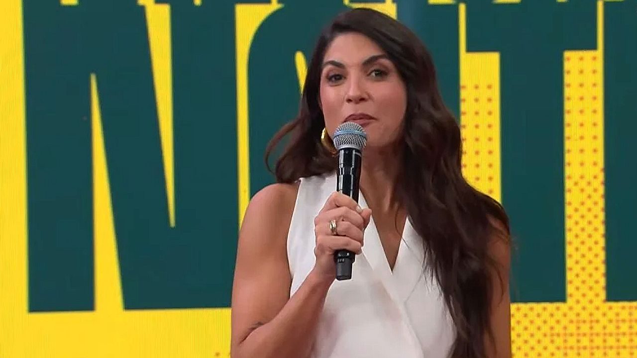 Apresentadora fala ao microfone em estúdio com telão colorido ao fundo durante programa de TV