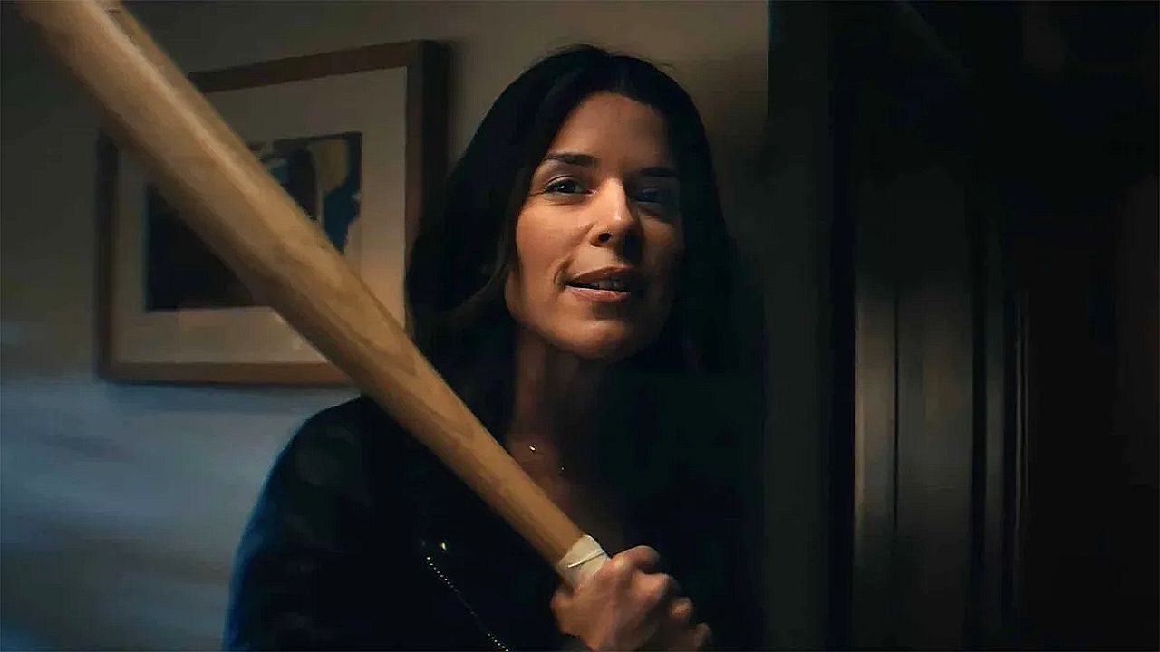 Uma mulher de cabelos longos e escuros aparece dentro de um ambiente interno, possivelmente um corredor ou sala de uma casa. Ela segura um taco de beisebol com as duas mãos, apoiado no ombro, enquanto olha à frente com expressão confiante ou desafiadora. A iluminação é baixa, criando sombras no ambiente. Ao fundo, há uma parede com um quadro pendurado e uma porta parcialmente visível.