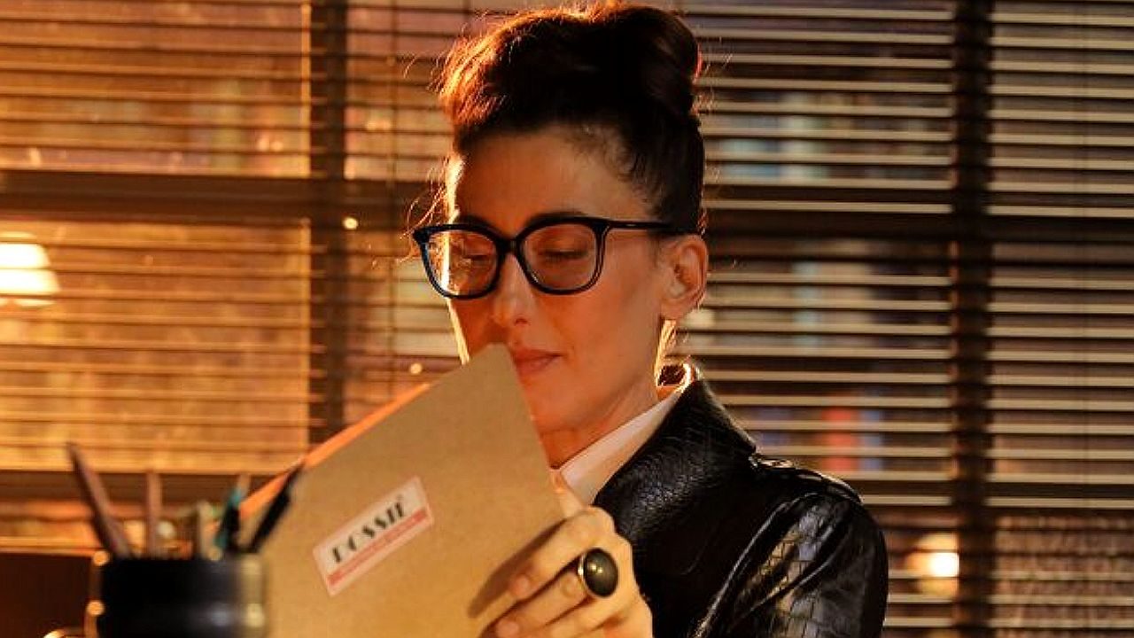 Mulher de óculos segura envelope com escrito dossiê em escritório com persianas ao fundo