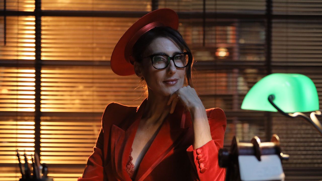 Paola Carosella caracterizada no programa Infiltrado na Cozinha, sentada à mesa com luminária verde ao lado