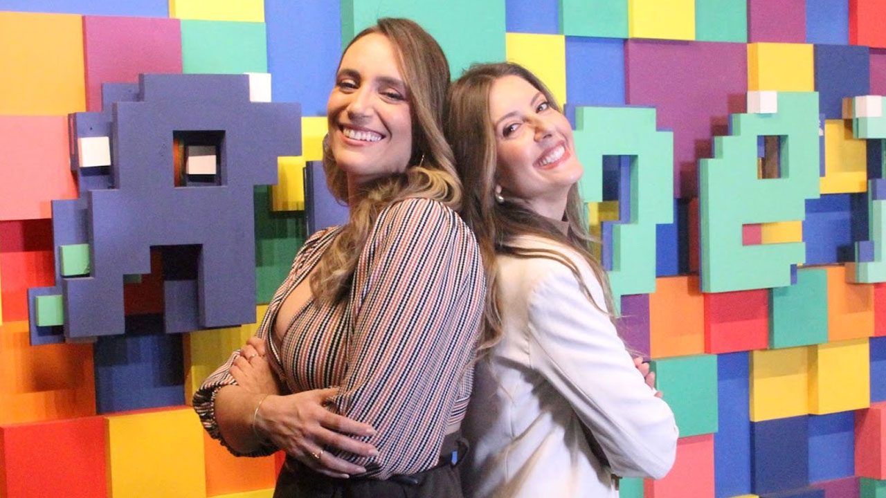 Paola Pretto e Taisa Pelosi sorrindo encostadas de costas diante do cenário colorido da RedeTV!