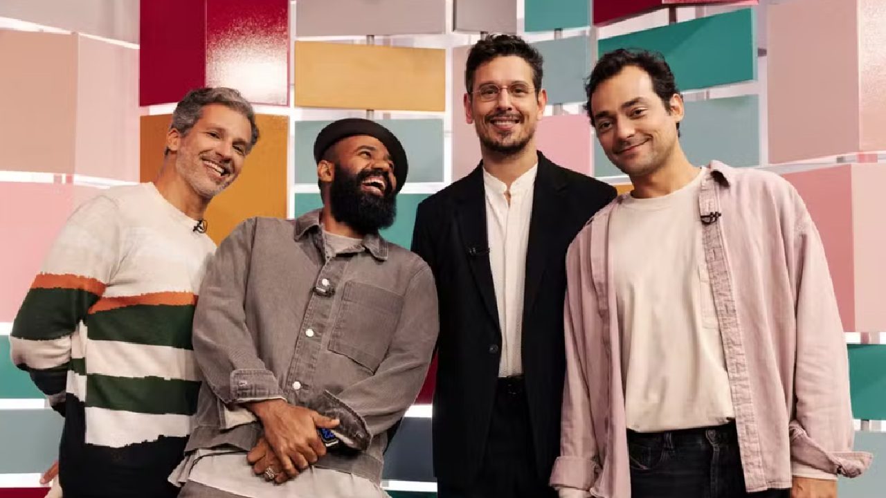 Quatro homens posam juntos sorrindo em frente a um painel colorido com blocos geométricos. O grupo transmite clima descontraído: um deles ri com a cabeça inclinada, enquanto os outros sorriem para a câmera.