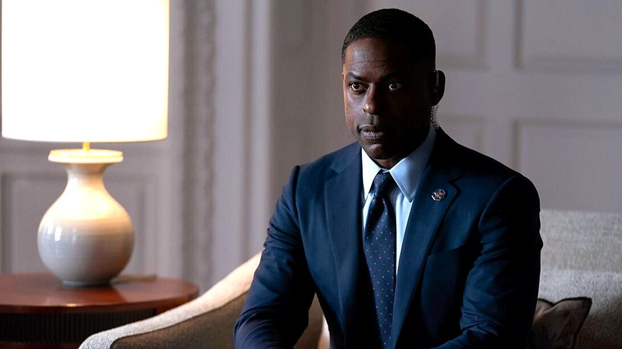 Homem de terno e gravata sentado em poltrona durante reunião em ambiente formal com abajur aceso ao lado