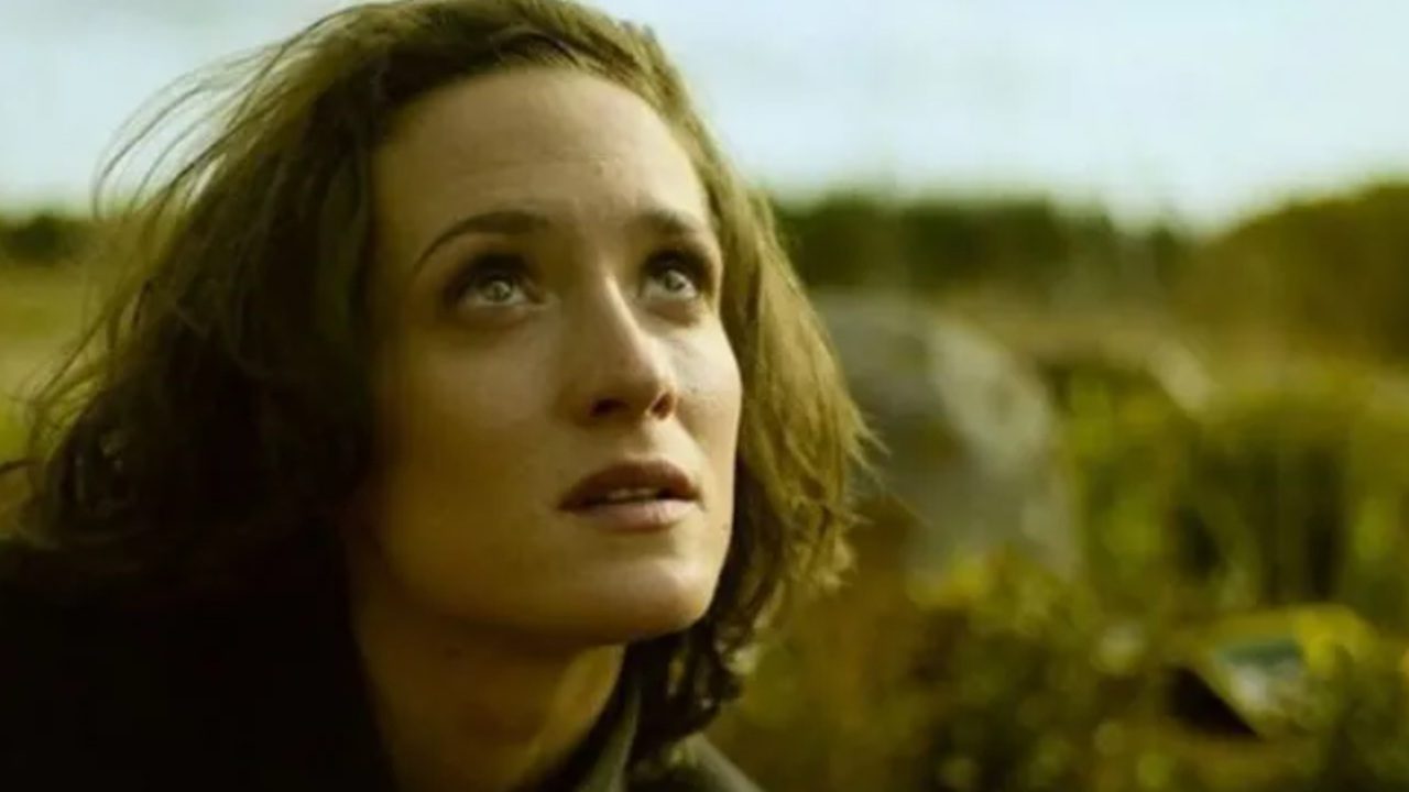 Mulher com expressão apreensiva olha para o alto em paisagem rural ao ar livre durante cena de filme ou série dramática