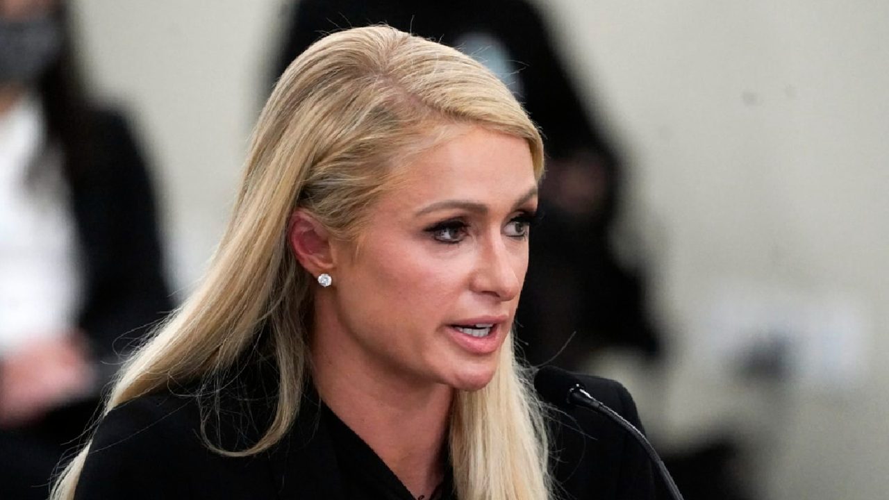 Paris Hilton falando ao microfone durante um evento formal, vestindo blazer preto e brincos de diamante, com expressão séria em ambiente institucional