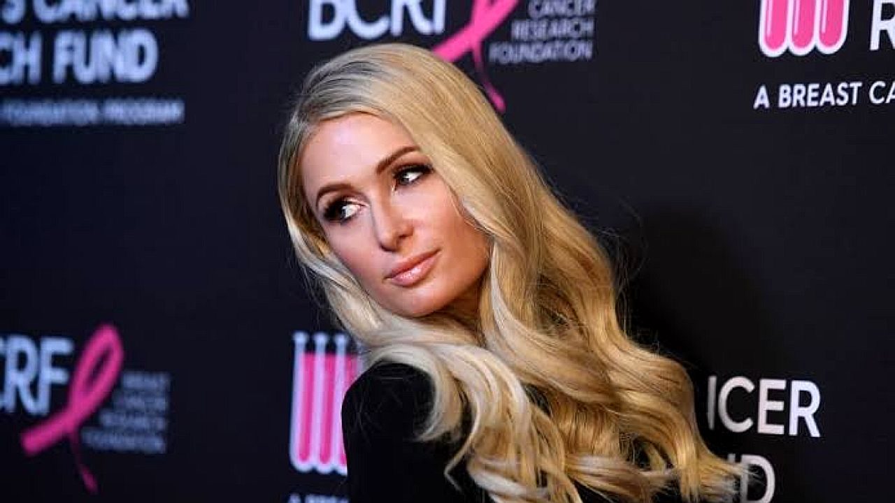 Paris Hilton posa com vestido preto e cabelos loiros em evento beneficente diante de painel de patrocinadores